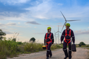 Male and female engineers in uniform, red safety bells, yellow hats, non-slip gloves for working at height walk into a wind turbine farm to perform maintenance. Clean energy from nature.