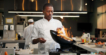 Black man, chef and fire with frying pan in kitchen at restaurant for fine dining, meal and creativity. Employee, professional and cook with food for culinary, hospitality and catering service