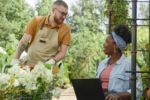 Positive Black Female Manager Using Laptop Chatting with Male Plant Nursery Worker in Garden