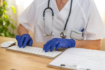 Doctor wearing blue nitrile gloves typing medical records on a computer keyboard while reviewing a patient chart in a medical office or clinic setting, ensuring accurate documentation